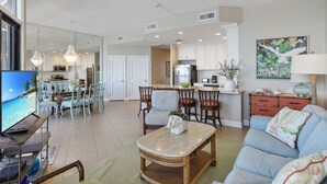 A spacious living area with a television, dining table, and chairs, a kitchen in the background, and a painting on the wall.