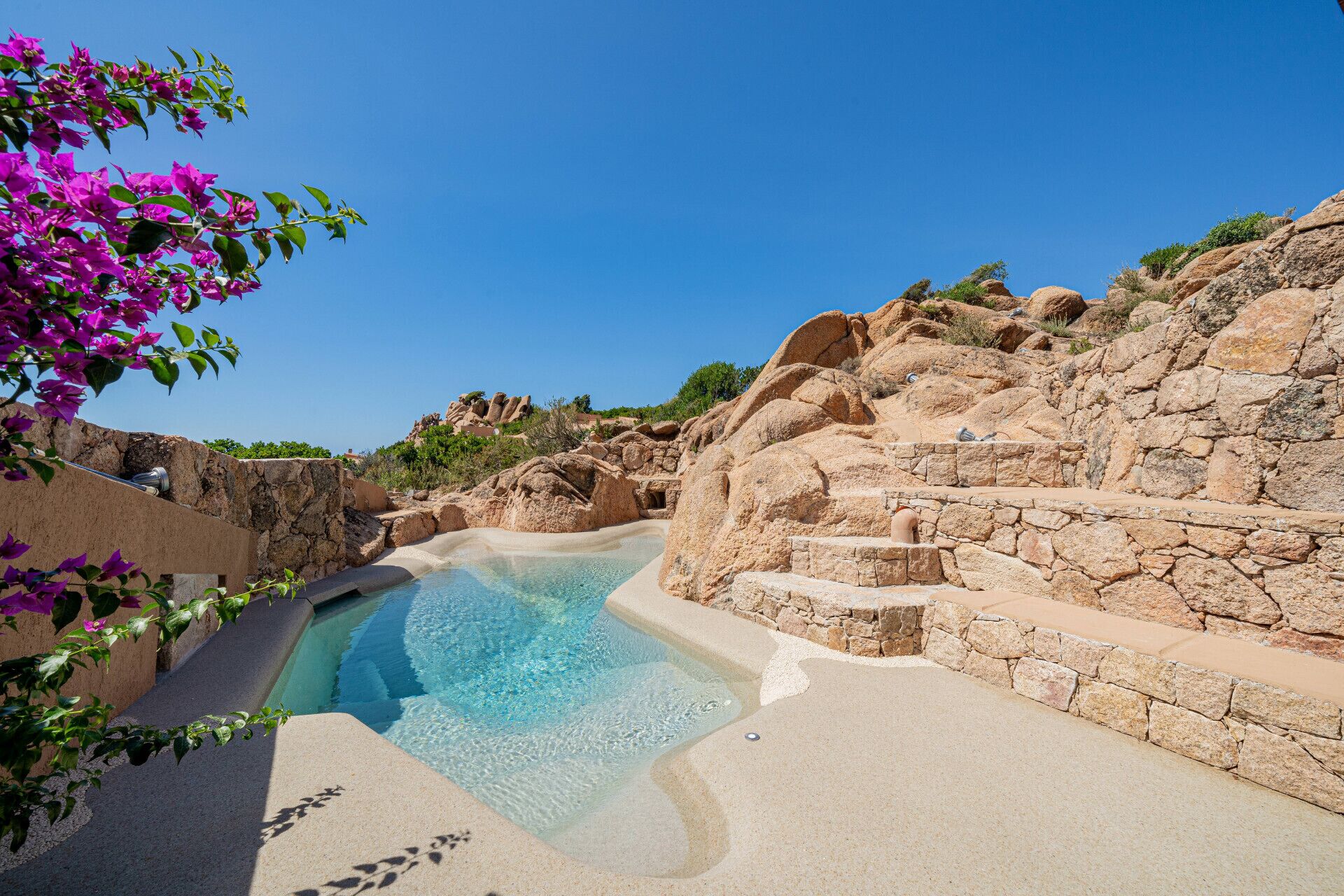 A clear blue pool surrounded by stone walls and steps, with purple flowers in the foreground.
