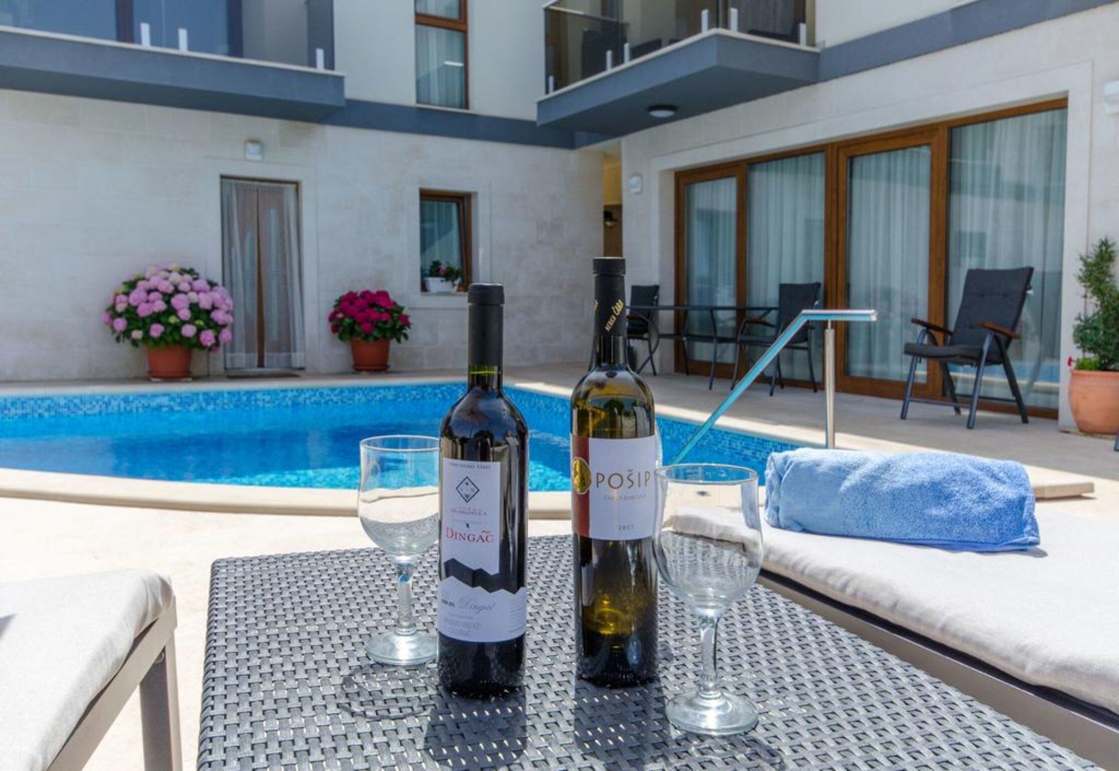 Zona junto a la piscina con dos botellas de vino y copas sobre una mesa, rodeada de sillones de descanso y una toalla azul.