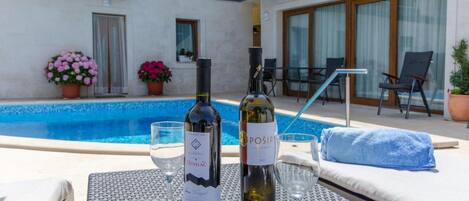 Zona junto a la piscina con dos botellas de vino y copas sobre una mesa, rodeada de sillones de descanso y una toalla azul.