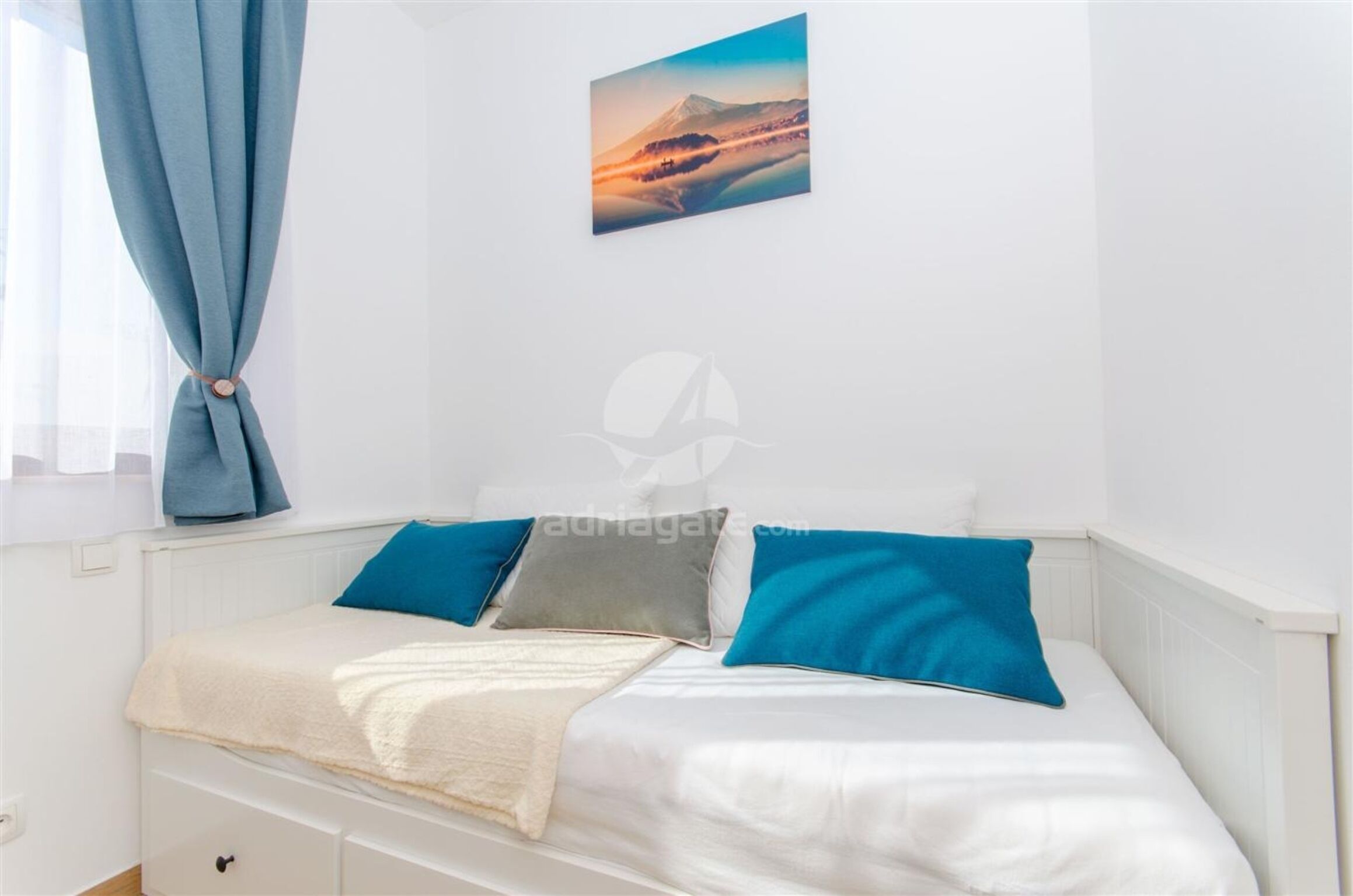 A bedroom with a bed, pillows, a blanket, a window with curtains, and a wall-mounted picture.
