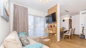 Una sala de estar moderna con un sofá, un televisor montado en la pared, una zona de comedor con sillas y una mesa, y un área de cocina con una planta.
