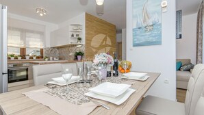 Una cocina moderna con horno integrado, un comedor con mesa de madera dispuesta para cuatro personas, y una sala de estar con sofá y un cuadro de veleros.