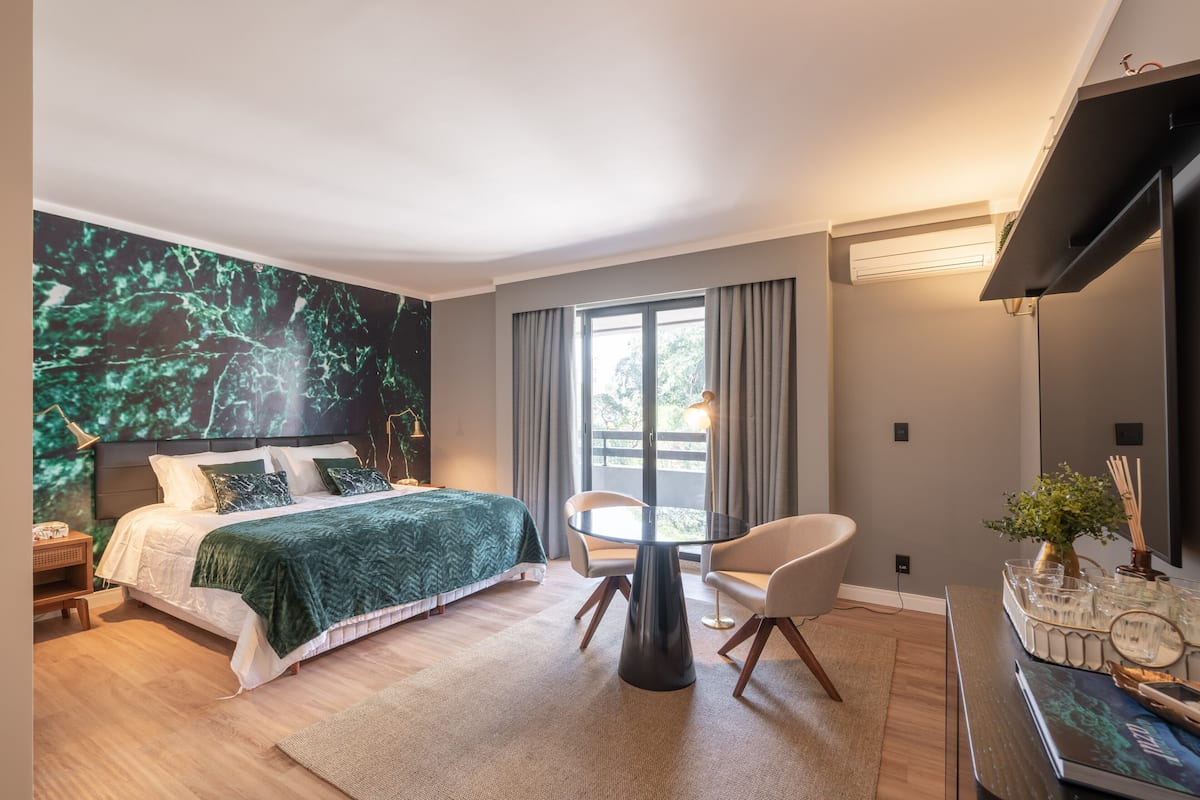 A modern bedroom with a bed, a round table, and a chair, a large window with curtains, and a wall-mounted TV.