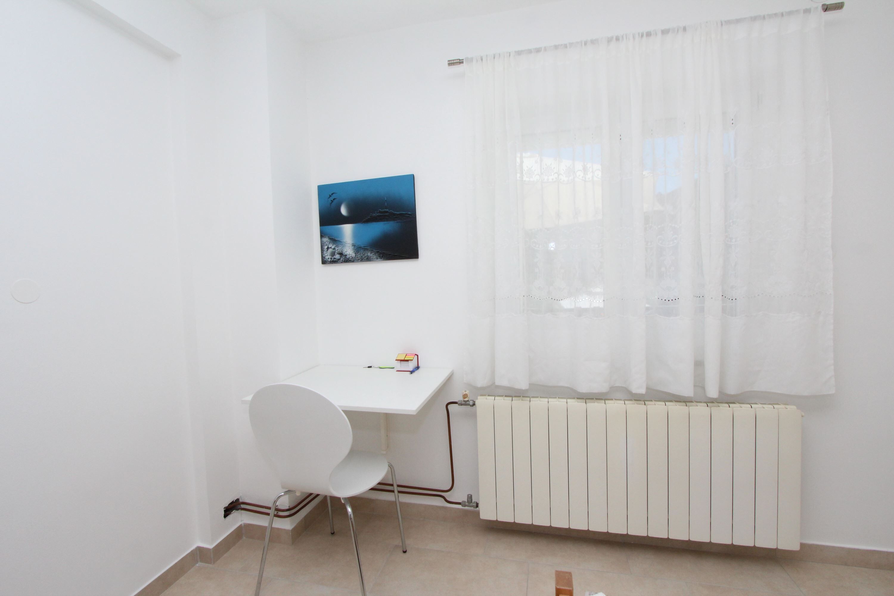 A room with a white desk, a chair, a radiator, and a window with sheer curtains.