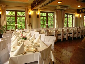 A formal dining room with tables set for a meal, large windows with curtains, and a ceiling fan.