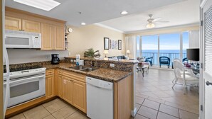 A kitchen with wooden cabinets, a microwave, a dishwasher, and a coffee maker. There is a dining area with a table and chairs, and a view of the ocean through large windows. - 16th-Floor Gulf-Front Condo at Boardwalk – Balcony Views, Resort Pool & Hot Tubs (Panama City Beach)