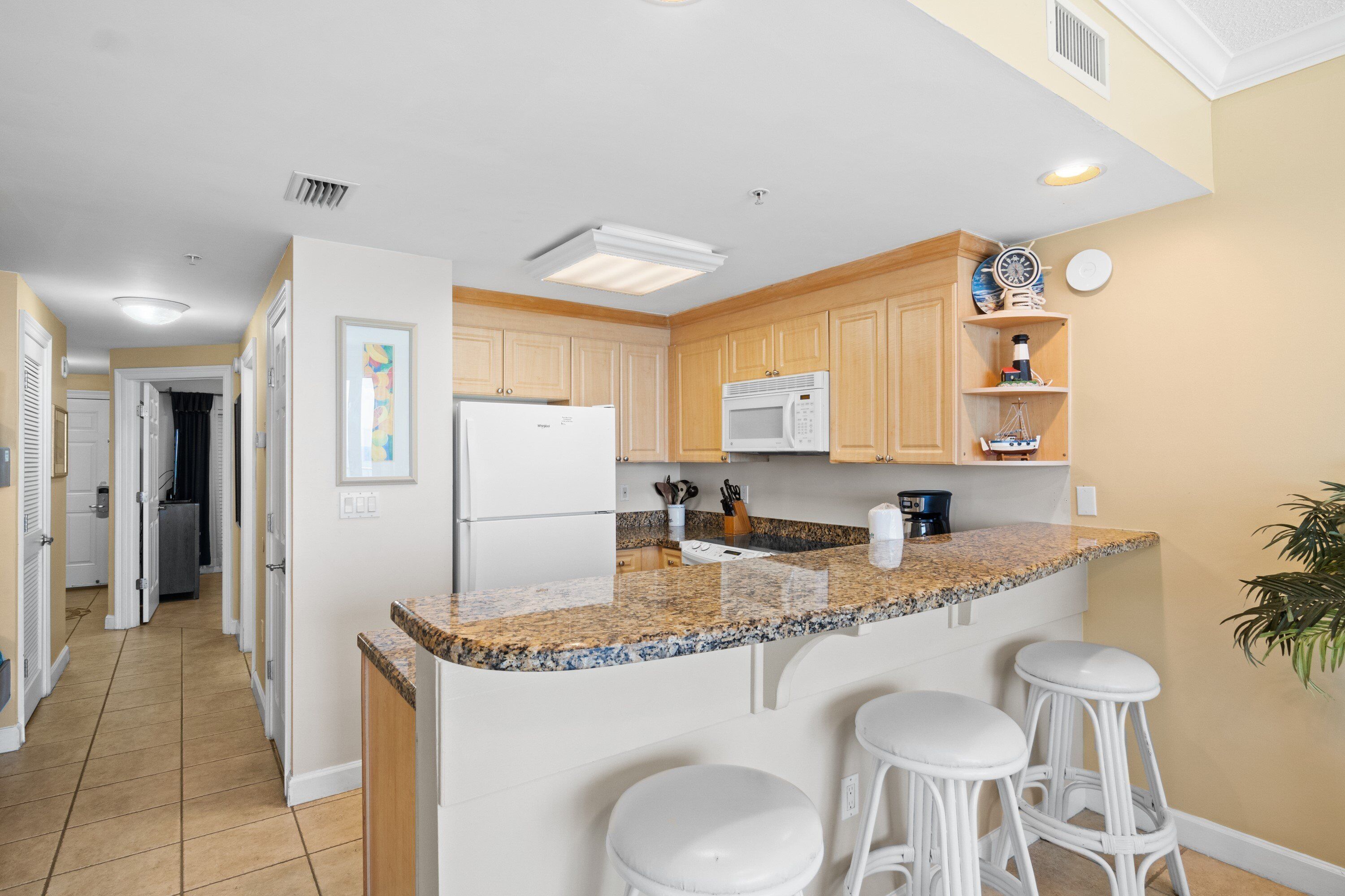 Cocina con encimera de granito, refrigerador blanco, microondas y armarios de madera. Hay tres taburetes de bar blancos y una planta en maceta.