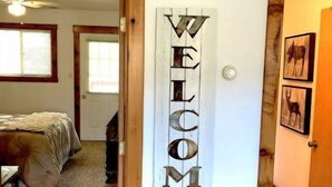 Un corridoio con un cartello che riporta la scritta 'WELCOME', un letto e una porta che dà su un'altra stanza.