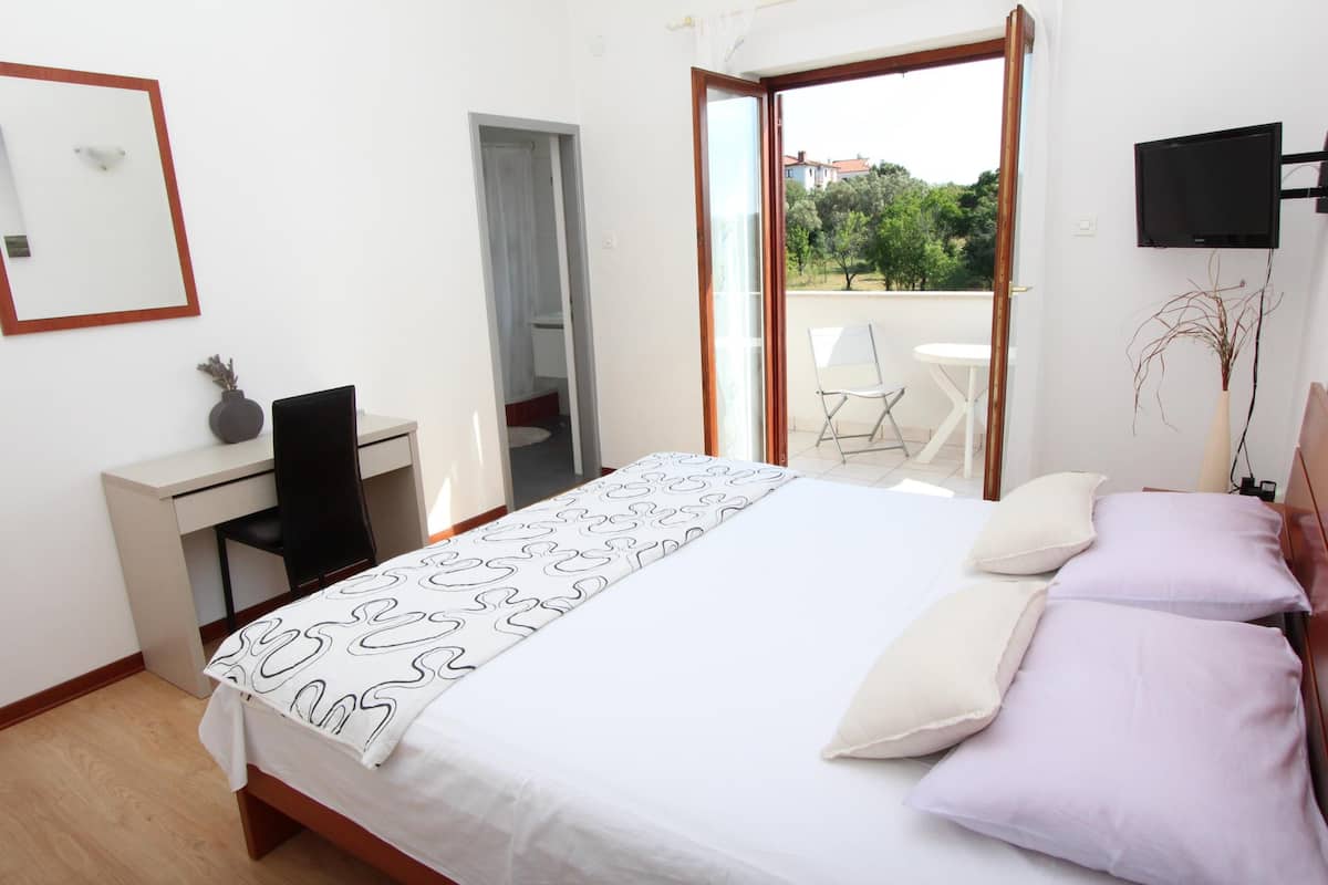 A bedroom with a bed, a desk, a chair, a TV, and a balcony with a view of greenery.