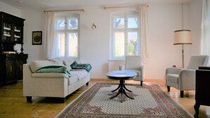 Een woonkamer met een bank, fauteuils, een bijzettafeltje, een kroonluchter en een groot raam met gordijnen.