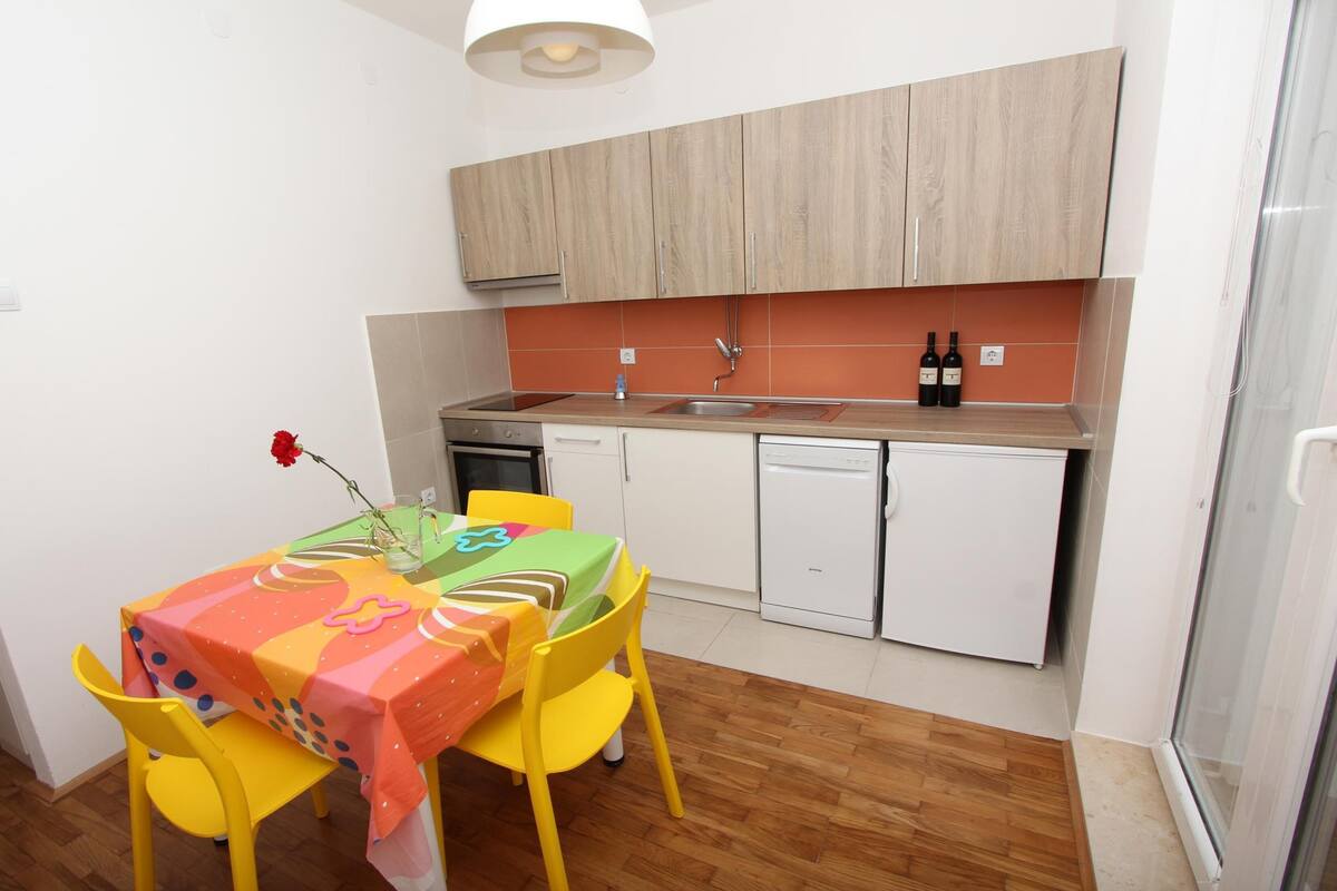 A small kitchen with wooden cabinets, a sink, and a dishwasher. There is a table with a colorful tablecloth and two yellow chairs.