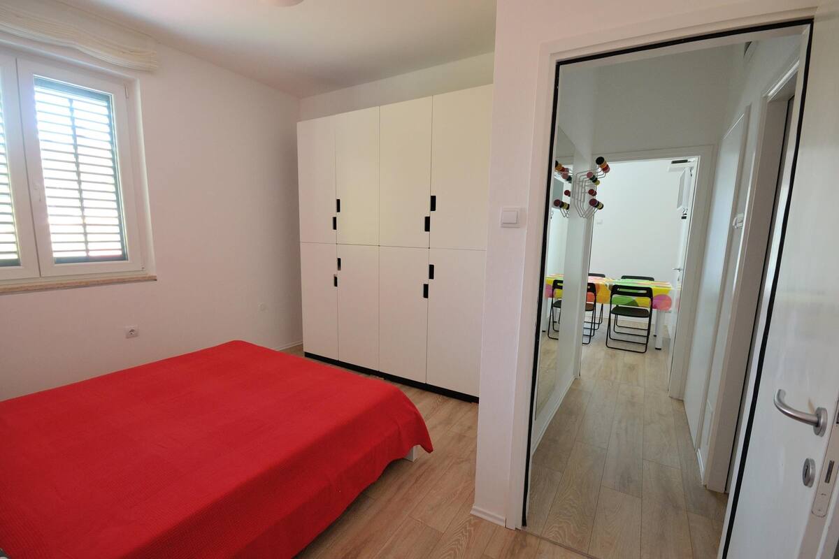 A bedroom with a red bedspread, white wardrobe, and a door leading to a dining area with chairs and a table.