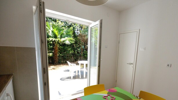 A room with a table and chairs, a door, and a view of a garden through an open door. - Apartments Paris (Rovinj)
