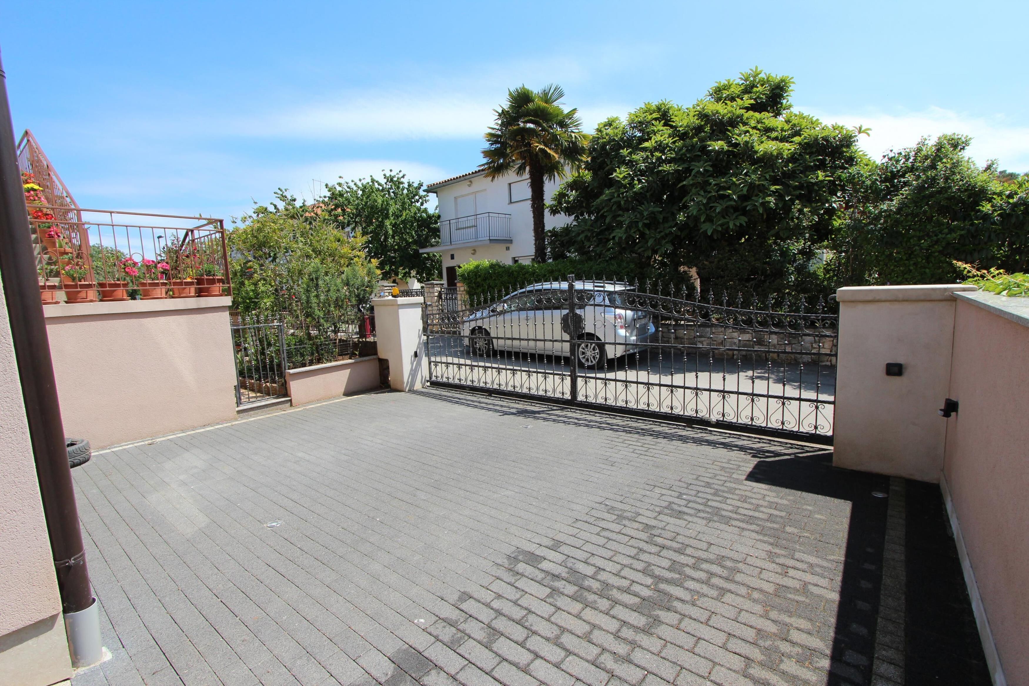Ein gepflasterter Außenbereich mit einem Metallzaun, einem weißen Auto im Inneren und einem Gebäude mit einem Balkon im Hintergrund.