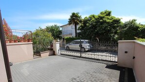Een verhard buitengebied met een metalen hek, een witte auto die er geparkeerd staat, en een gebouw met een balkon op de achtergrond.