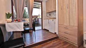 Een kleine keuken met houten kastjes, een eethoek met een tafel en stoelen, en een uitzicht door een deur op een buitenruimte.