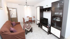 Una sala de estar con un sofá marrón, un televisor, un mueble de vidrio y un comedor con una mesa y sillas.