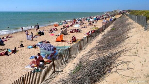 A beach with people sunbathing and swimming, a wooden fence, and a clear sky.