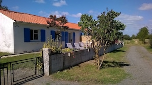 A house with a red-tiled roof, a white exterior, and blue shutters. There is a small garden with a tree and a concrete wall with a gate.