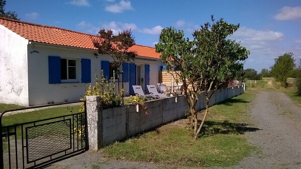 A house with a red-tiled roof, a white exterior, and blue shutters. There is a small garden with a tree and a concrete wall with a gate.