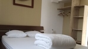 A neatly made bed with white linens and pillows, a wooden headboard, a framed picture on the wall, and a wooden wardrobe with hanging space.