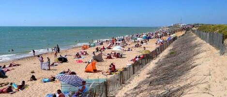 A beach with people sunbathing and swimming, a wooden fence, and a clear sky.