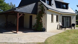 Une maison à deux étages avec un porche couvert, un vélo garé à l’extérieur et une chaise sur la terrasse.