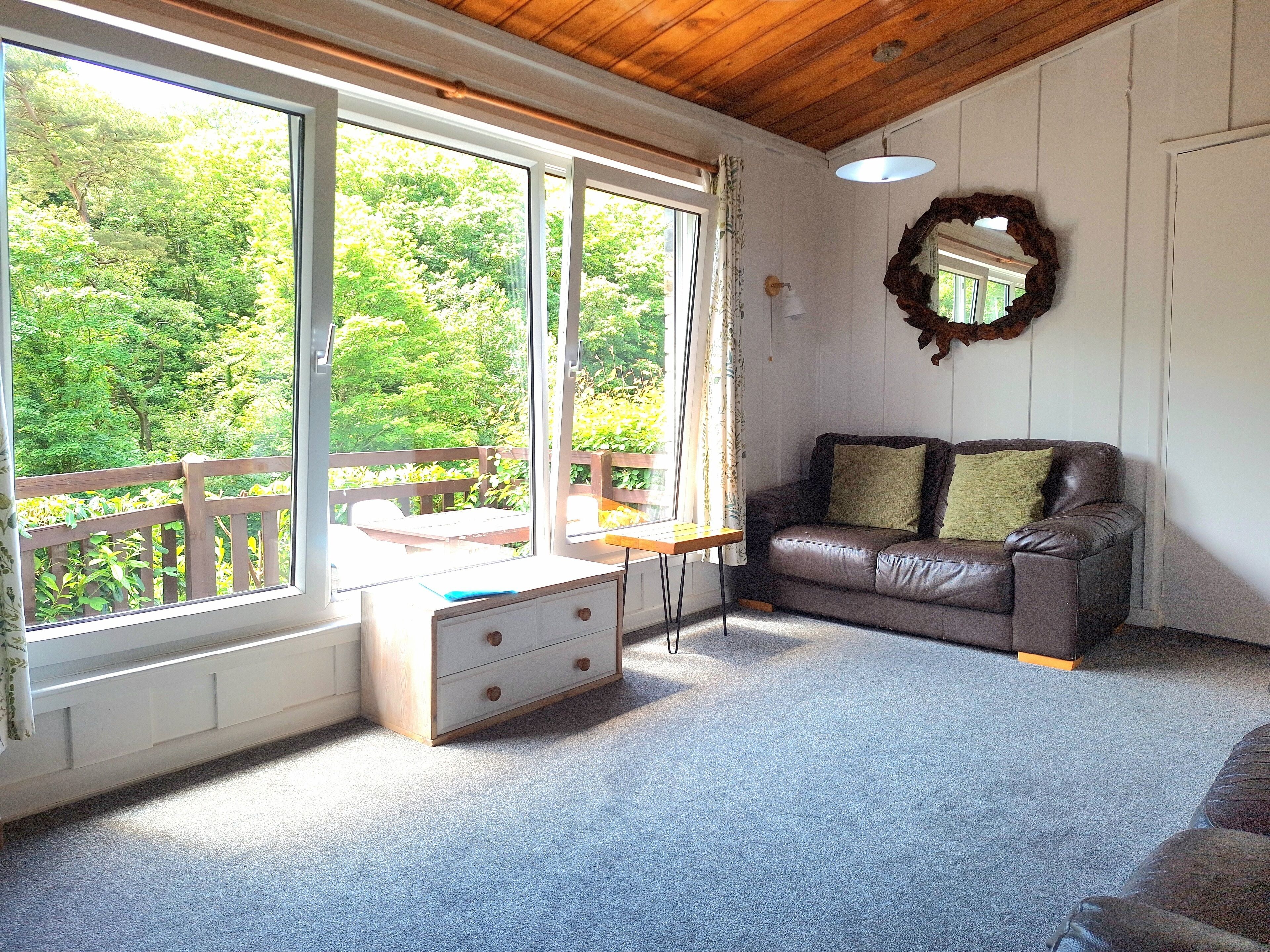 A cozy living room with a leather sofa, a wooden table, and a large window offering a view of greenery.