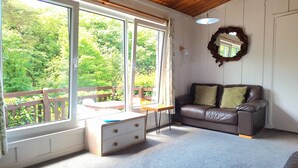 A cozy living room with a leather sofa, a wooden table, and a large window offering a view of greenery. - Parada Cottage@Mineshop Holiday Cottages, a woodland stroll to Crackington Haven (Bude)
