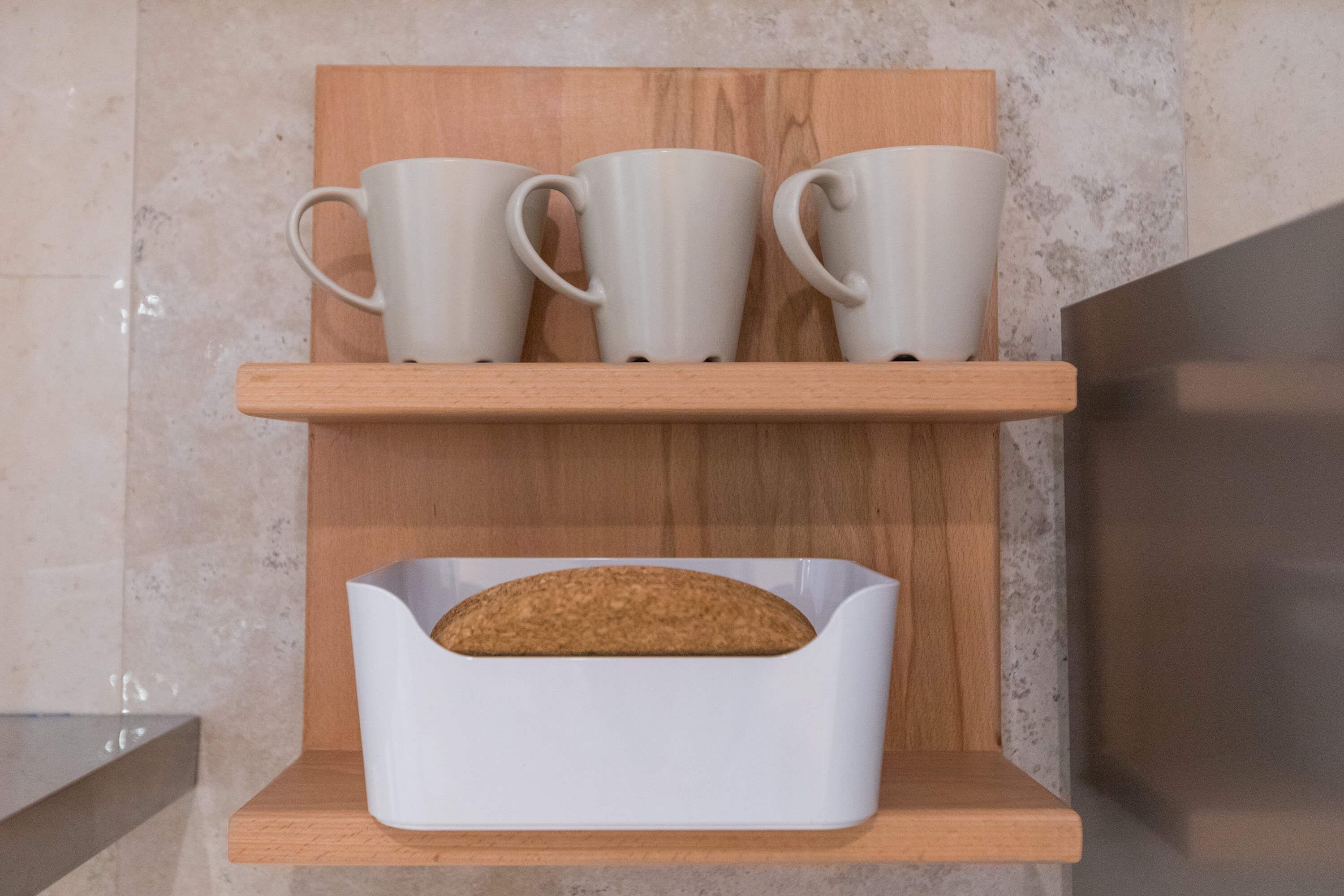 Una estantería de cocina con tres tazas blancas y una barra de pan en un panera blanco.