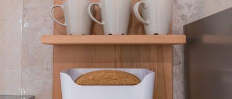 Una estantería de cocina con tres tazas blancas y una barra de pan en un panera blanco.