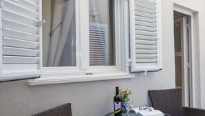 A balcony with wicker chairs, a table with a bottle of wine, and a window with white shutters. - Apartment Dam (Dubrovnik)