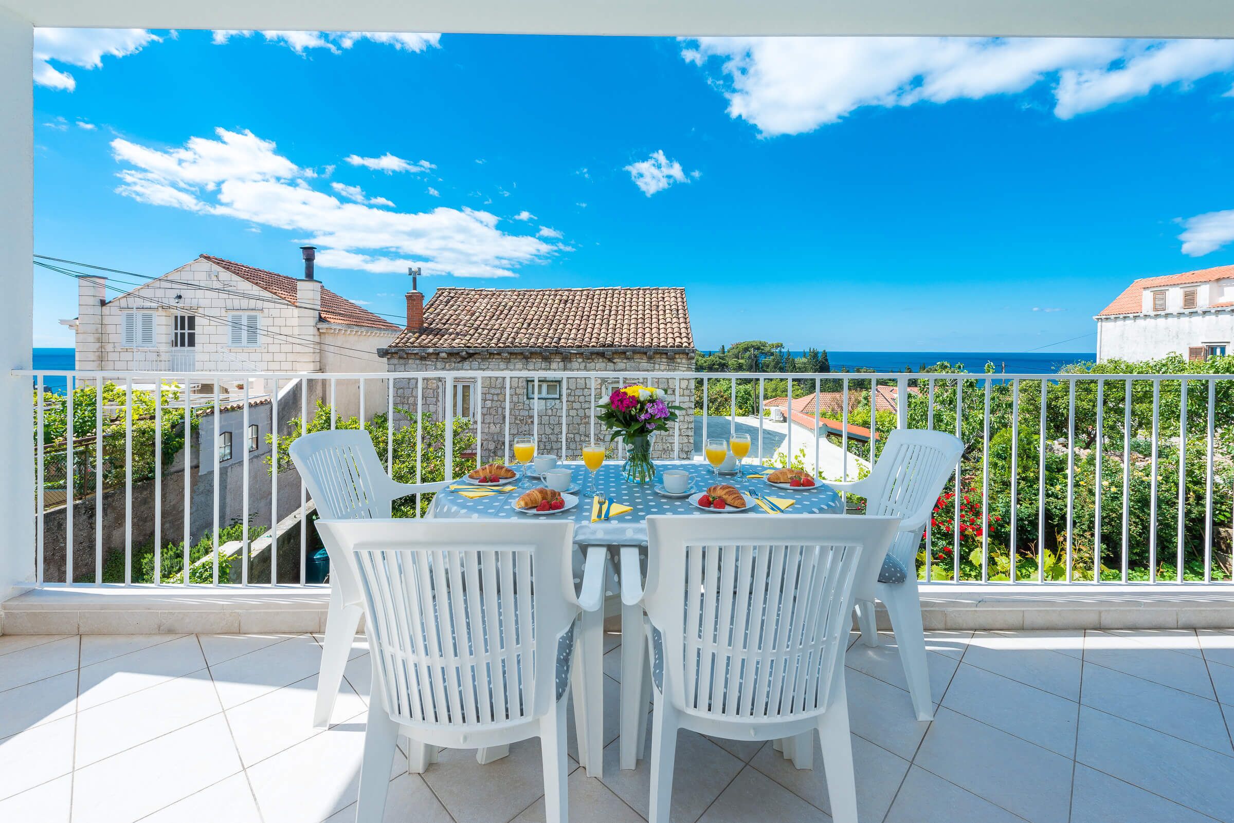 A balcony with a table set for breakfast, white chairs, and a view of the sea and buildings.