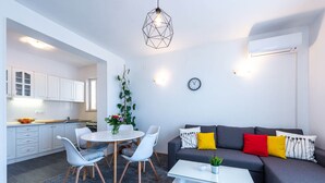 Una sala de estar moderna con comedor, un sofá gris con cojines de colores, una mesa de centro blanca y una lámpara colgante de diseño geométrico.