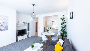 Una sala de estar moderna con un televisor de pantalla plana, un comedor con mesa redonda y sillas, y una cocina al fondo.