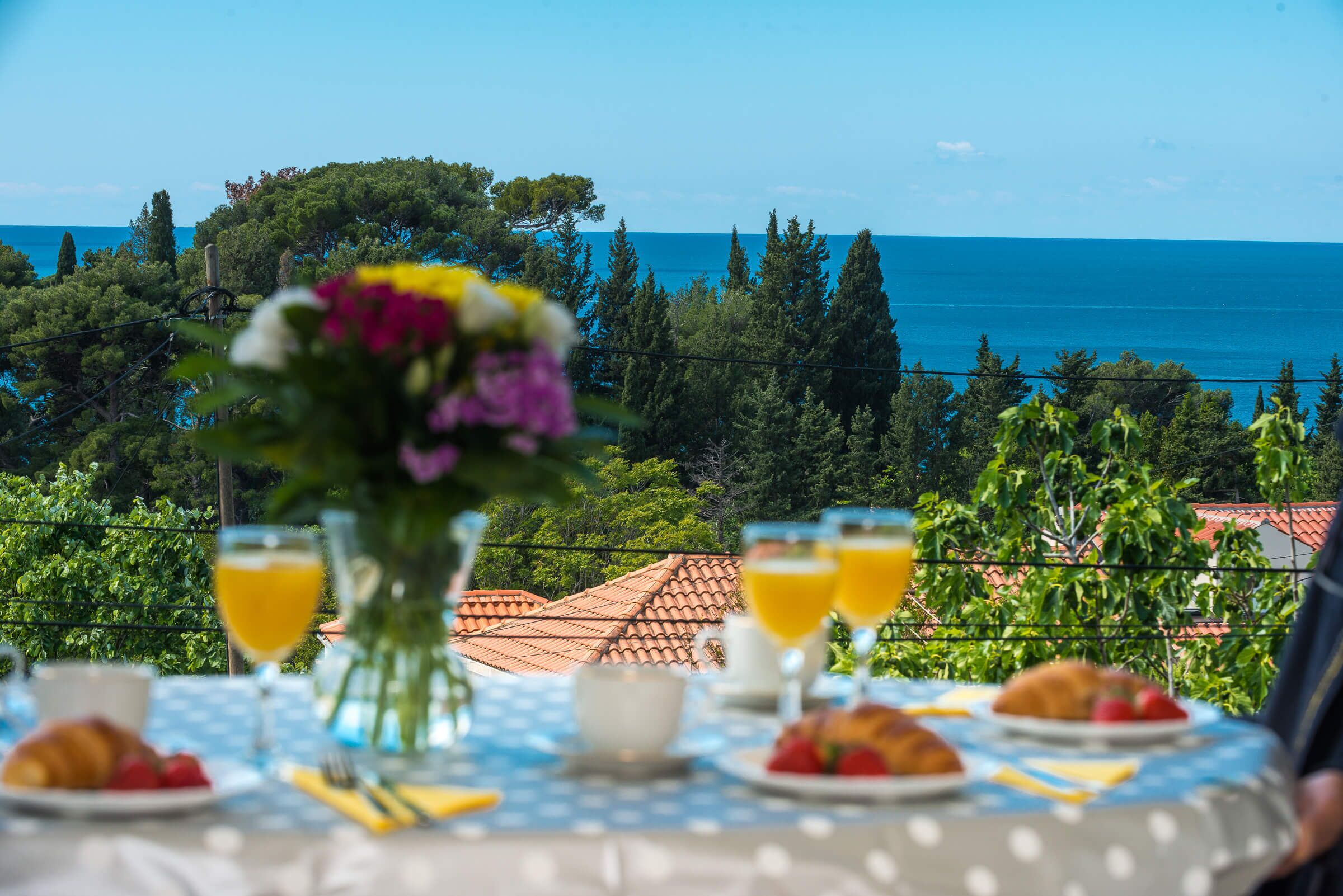 Ett dukat bord för frukost med utsikt över havet, en vas med blommor och en croissant.