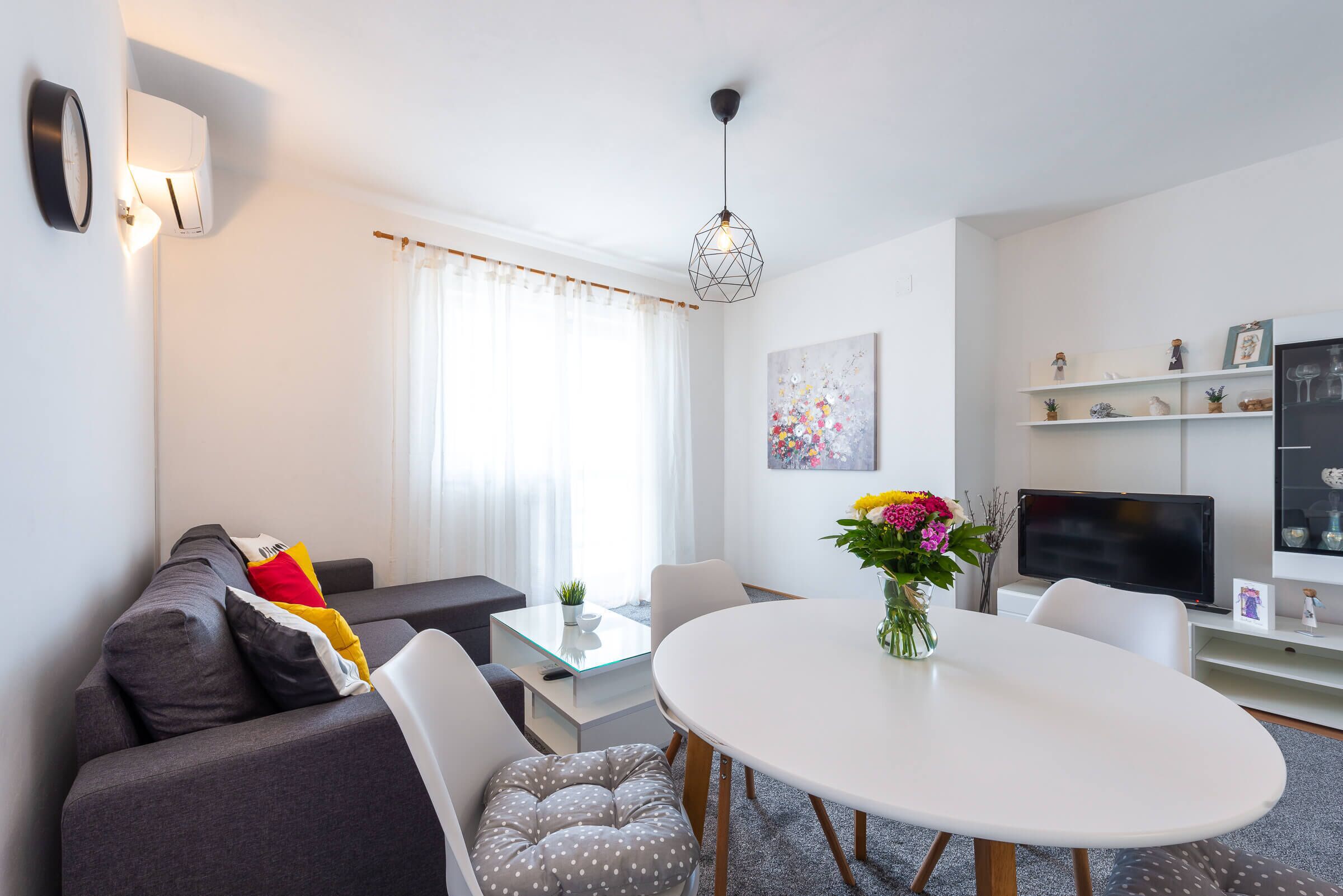 A modern living room with a grey sofa, white chairs, a glass coffee table, a white dining table, a TV, and a decorative vase with flowers.