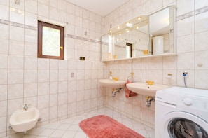 Un baño con bidé, dos lavamanos, un espejo con luces, una ventana y una lavadora.