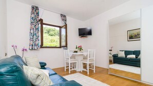Una sala de estar con un sofá de cuero verde, sillas blancas, una mesita con un florero y un televisor de pantalla plana montado en la pared. Se ve un espejo grande que refleja un sofá verde y un cuadro.