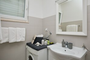 A bathroom with a washing machine, a sink, and a mirror. There are towels hanging on the wall and toiletries on the sink counter. - Bahari Apartment (Dubrovnik)