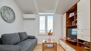 Un salon moderne avec un canapé gris, une table basse en bois agrémentée de livres et d’un vase, un téléviseur posé sur un meuble en bois, et une grande horloge au mur.