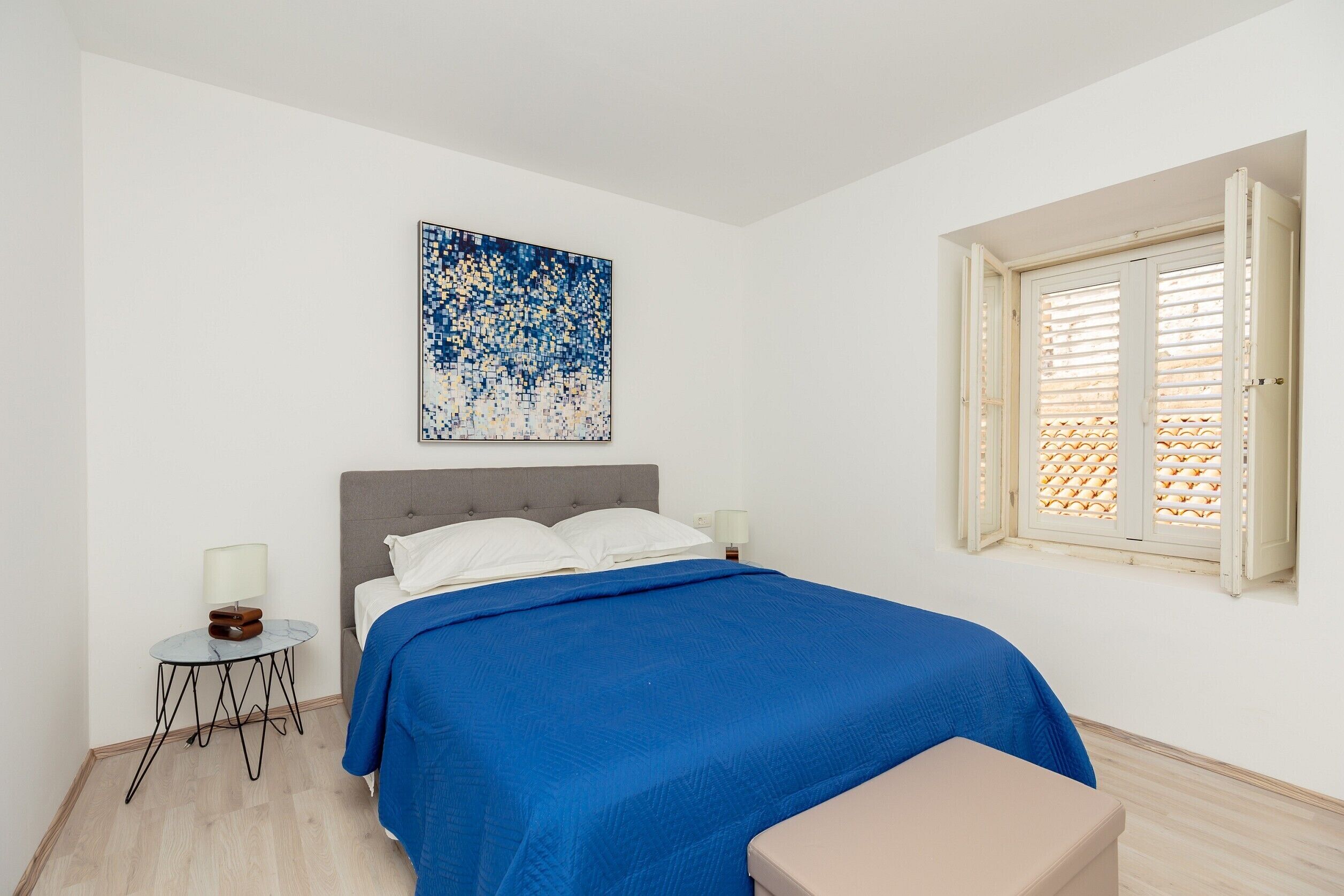 A bedroom with a bed, a bedside table with a lamp, a window with shutters, and a wall-mounted artwork.