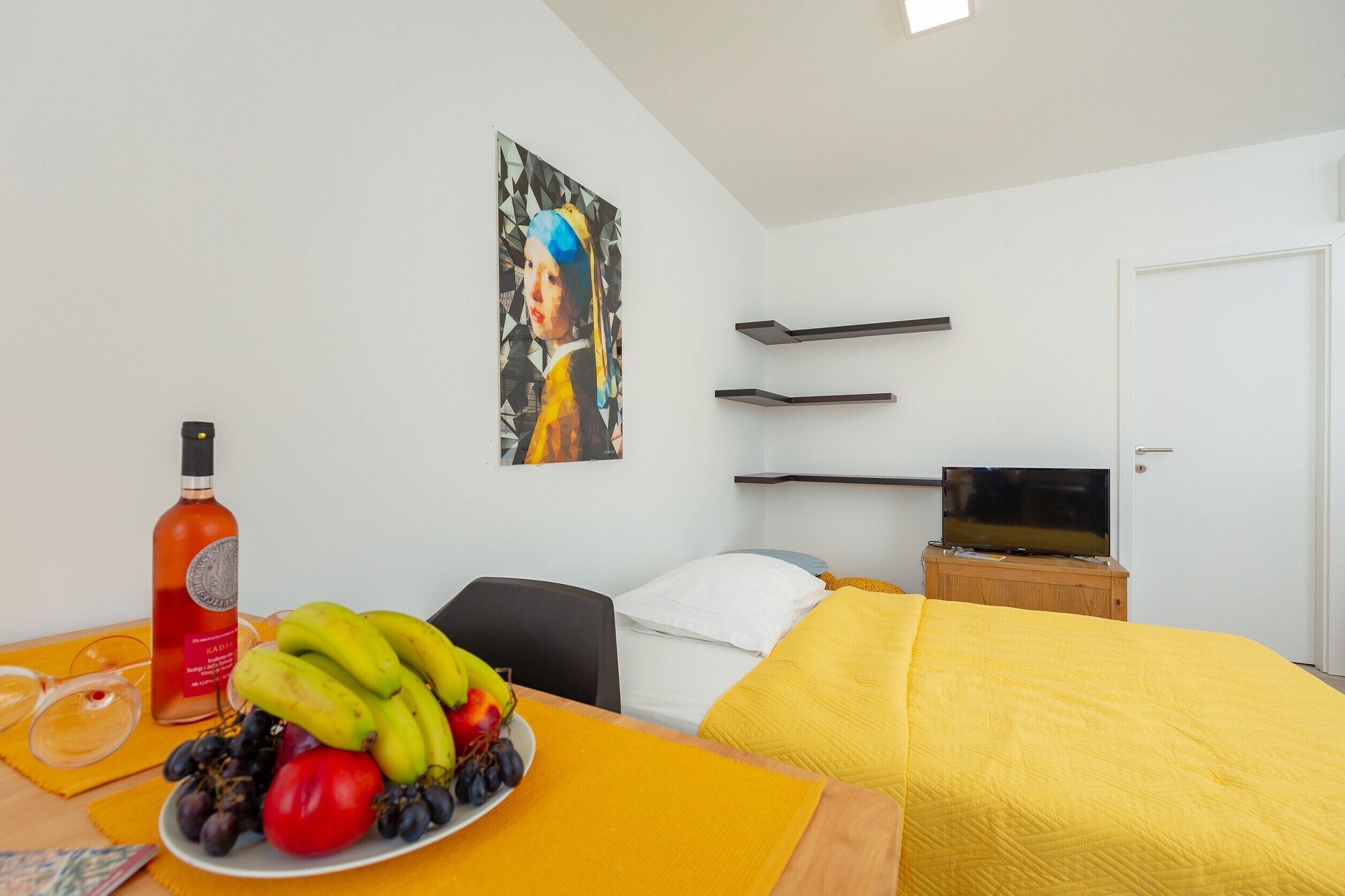 Quarto de hotel com cama, televisão, pintura, garrafa de bebida alcoólica e prato de frutas.