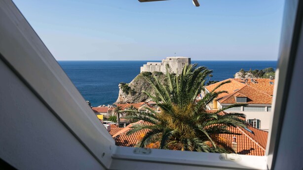 Blick aufs Meer vom Fenster aus, mit Blick auf ein historisches Gebäude auf einer Klippe, im Vordergrund Palmen und Gebäude mit Terrakotta-Dächern.