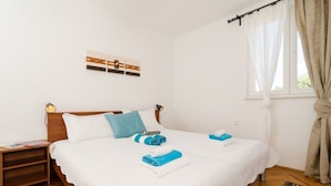 A neatly made bed with white bedding and blue towels, a wooden nightstand with books, a wall-mounted light, and a window with curtains.