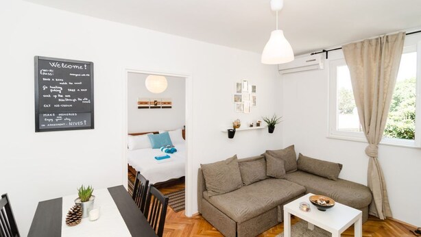 A modern living room with a sofa, a dining table with chairs, a hanging light fixture, and a window with curtains.
