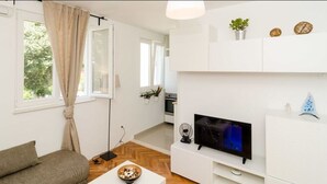 A modern living room with a sofa, a flat-screen TV, a dining table with a bowl, and a kitchen in the background.