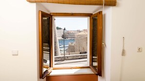 A room with a window overlooking a historic cityscape, including a fortified structure and a body of water. - Studios Lončarica (Dubrovnik)
