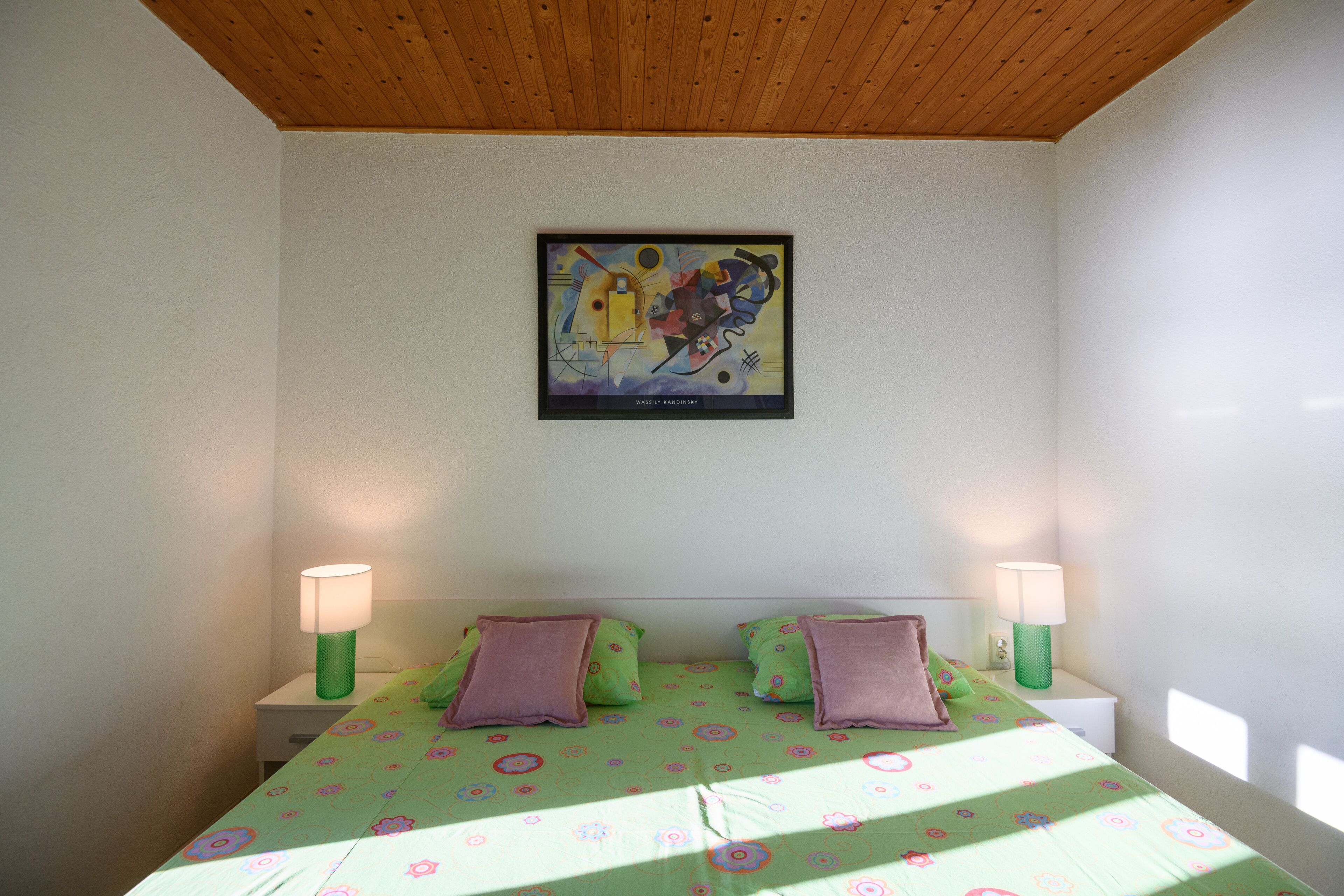 Ein Schlafzimmer mit Holzboden, einem Bett mit grüner Bettwäsche und violetten Kissen, zwei Nachttischlampen und einem gerahmten abstrakten Gemälde an der Wand.